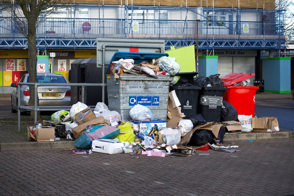 An outdoor urban scene showing a large pile of mixed rubbish and waste materials situated on a paved area adjacent to a railing and a silver car with a yellow and white license plate. The waste includ