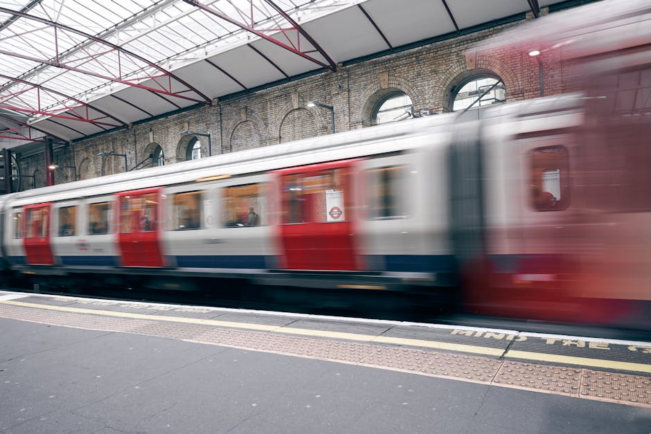 A motion-blurred photograph of a red and white London Underground train arriving or departing from Highbury and Islington station. The train is seen moving swiftly across the platform, which has a tex