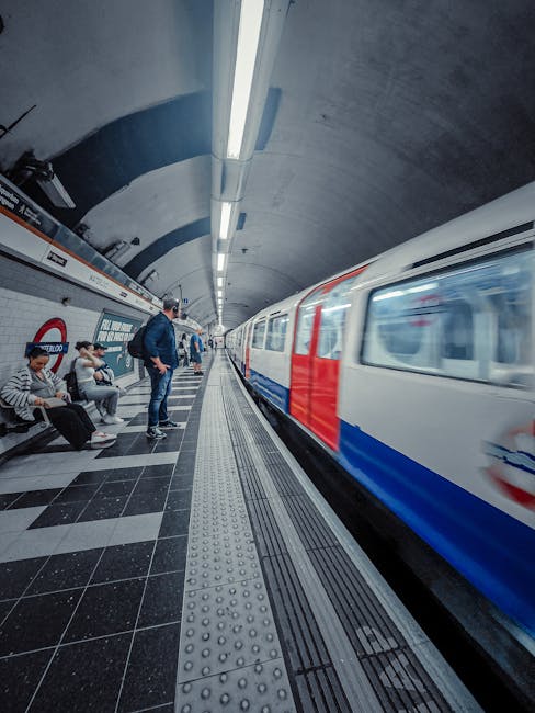 A motion-blurred photograph of a red and white London Underground train arriving or departing from Highbury and Islington station. The train is seen moving swiftly across the platform, which has a tex